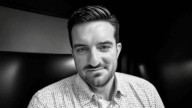 A man with short hair wearing a checkered shirt, smiling in a dimly lit space