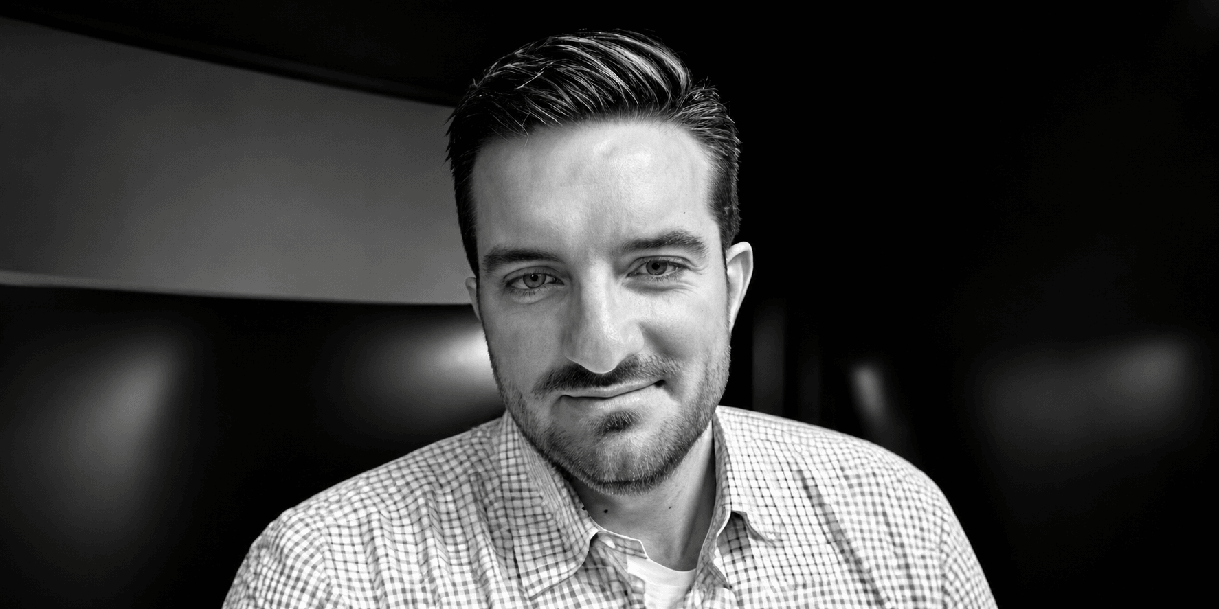A man with short hair wearing a checkered shirt, smiling in a dimly lit space