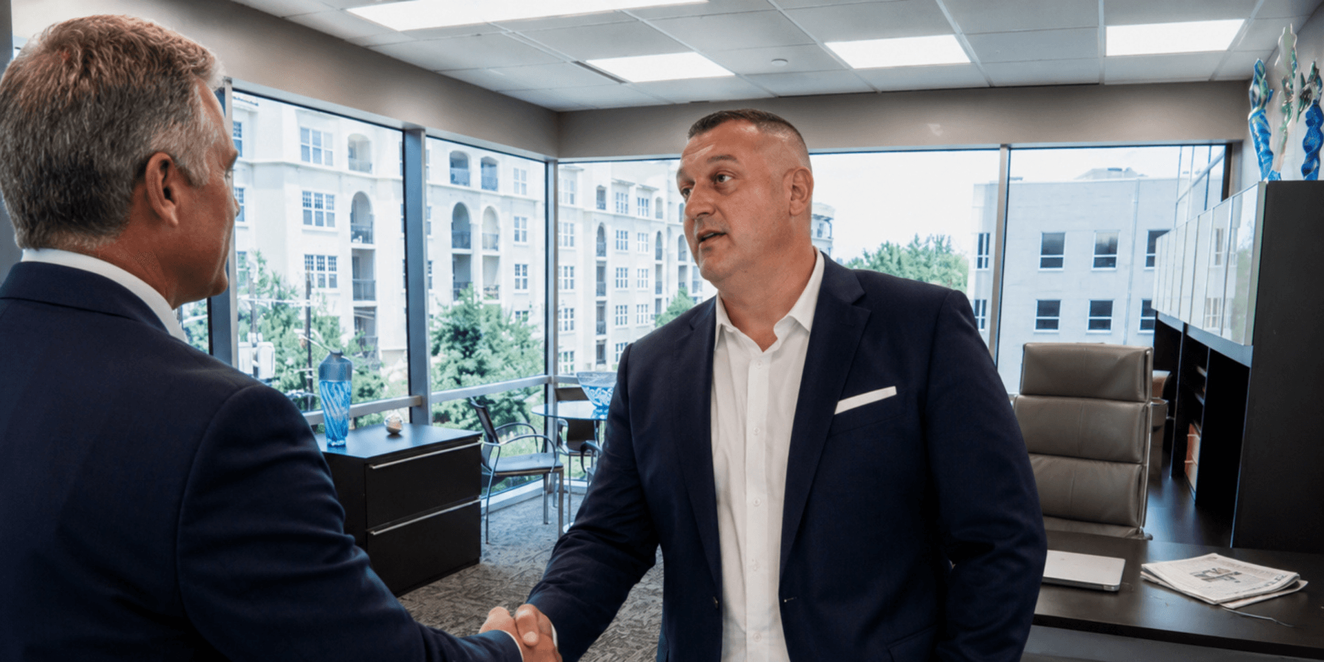 Two men shake hands in a modern office with large windows and a view.