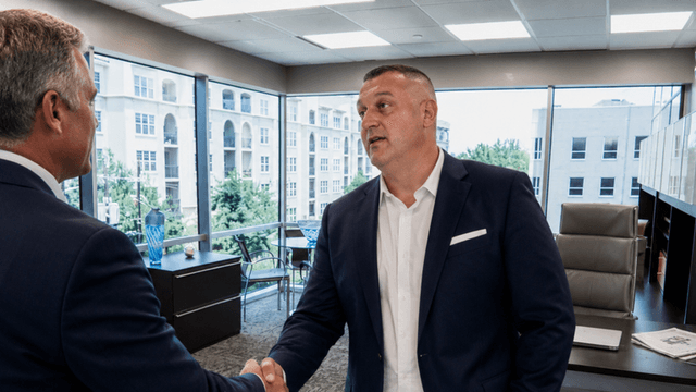 Two men shake hands in a modern office with large windows and a view.