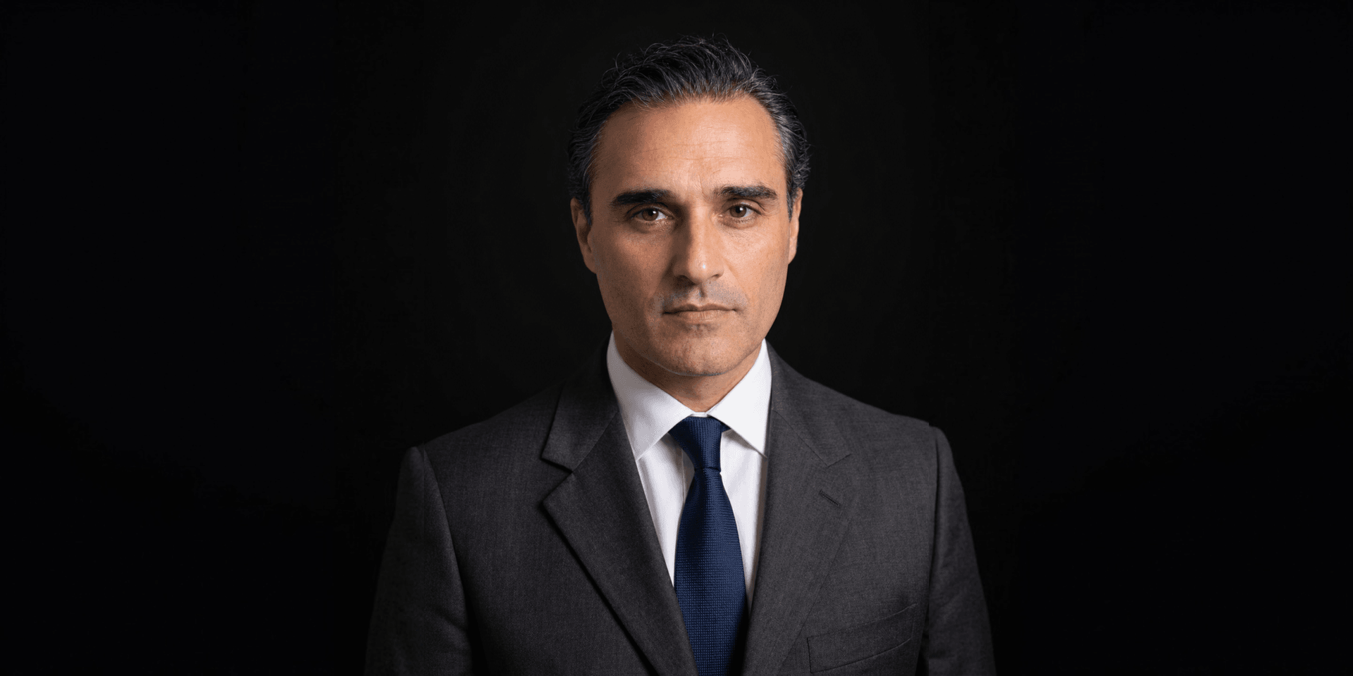A serious man in a dark suit and tie against a black background
