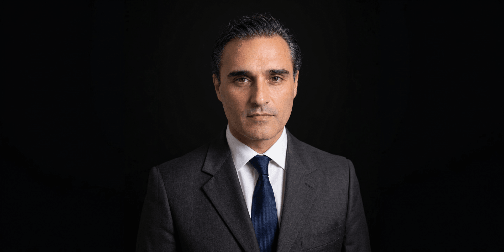 A serious man in a dark suit and tie against a black background