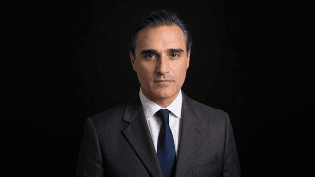 A serious man in a dark suit and tie against a black background