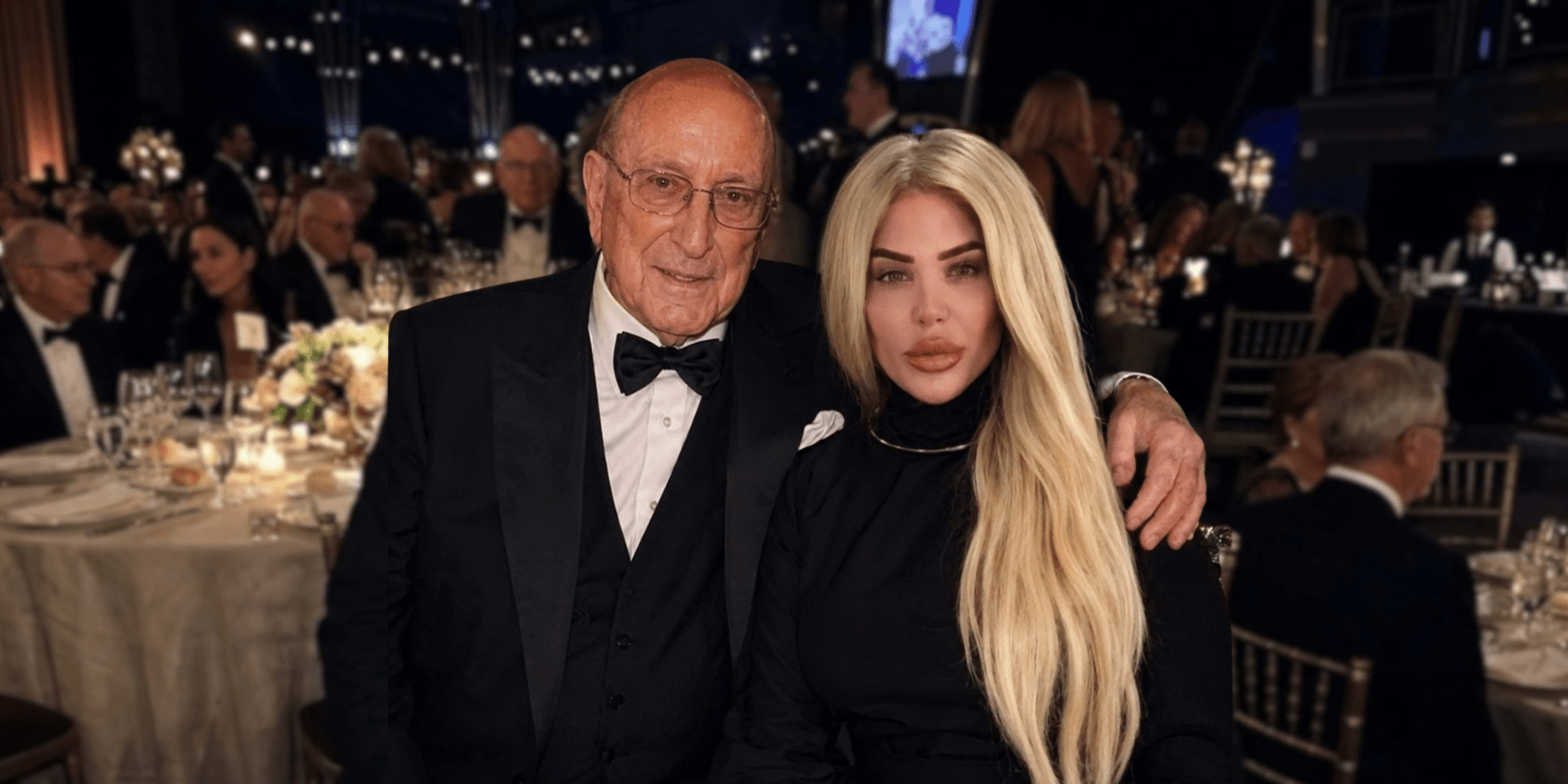 An elderly man and a woman with long blonde hair pose together at a formal event