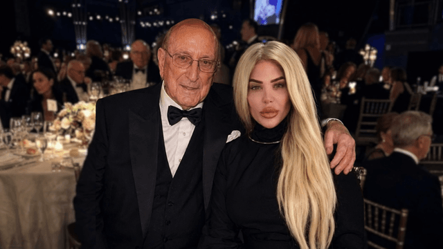 An elderly man and a woman with long blonde hair pose together at a formal event