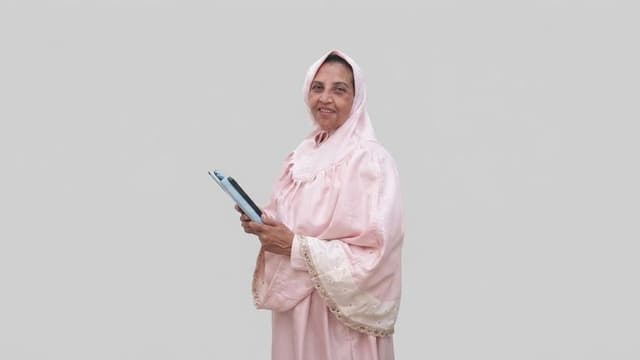 An older woman wearing a pink dress and headscarf holds a tablet and smiles.