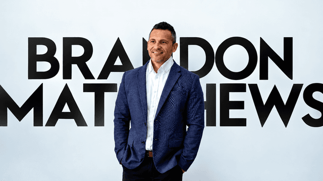 Smiling man in a blazer stands in front of a bold name backdrop