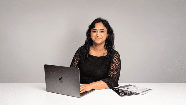 A woman with black curly hair sits at a desk with a laptop and notebooks