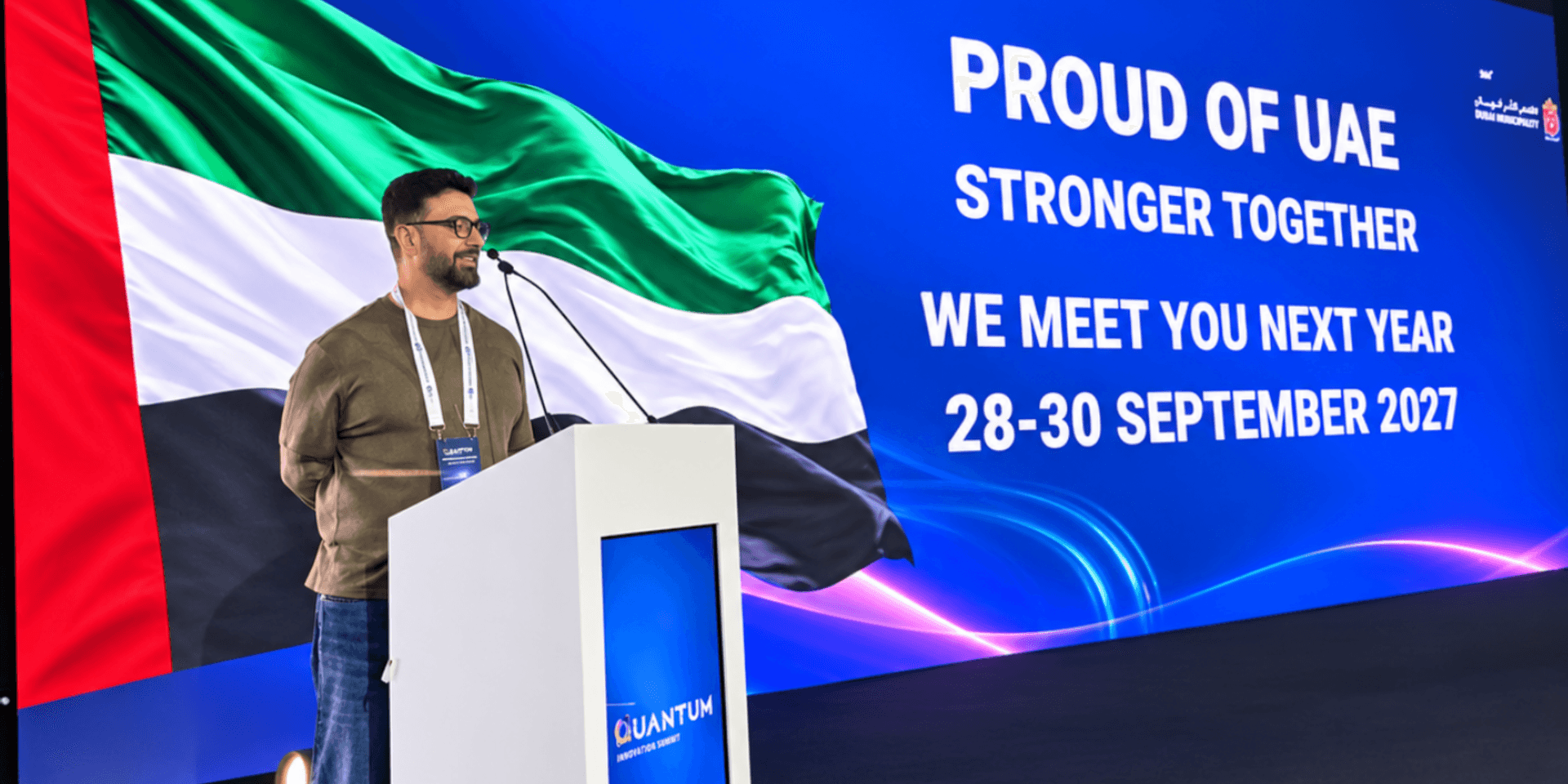 A speaker addresses an audience with a UAE flag backdrop and event details displayed.