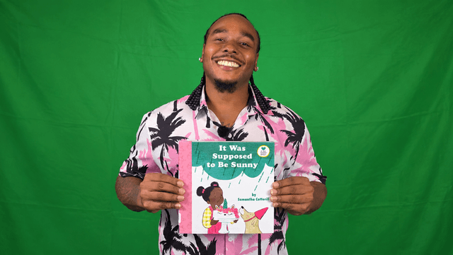 Smiling man in a floral shirt holding a children's book against a green background