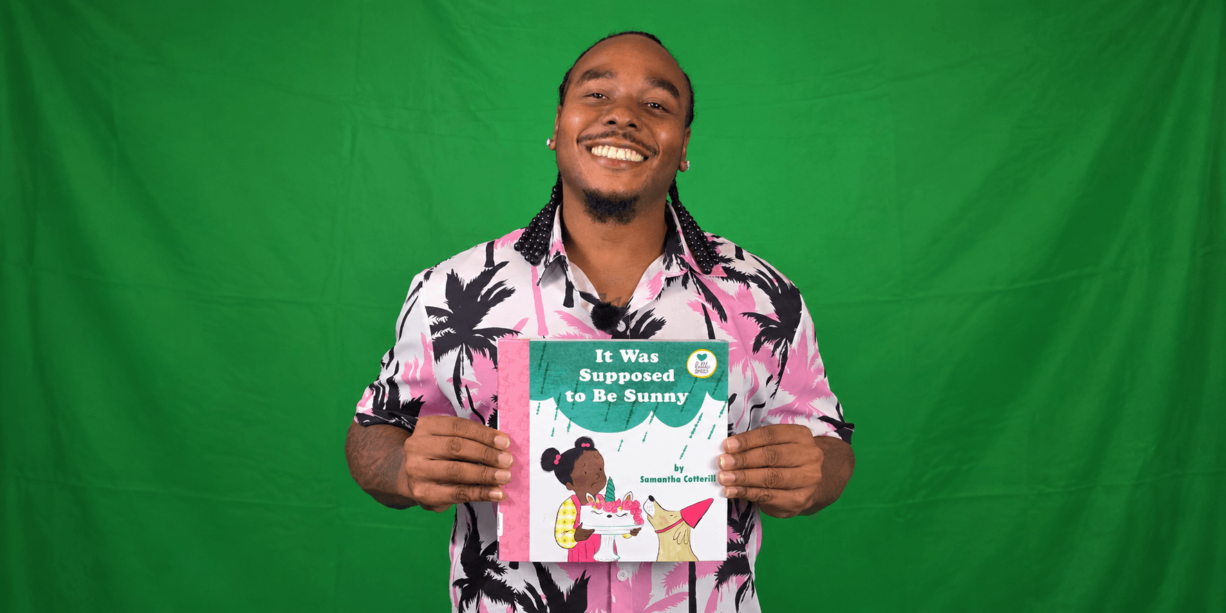 Smiling man in a floral shirt holding a children's book against a green background