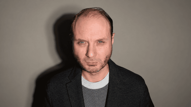 A man with short hair and a serious expression, wearing a dark jacket.