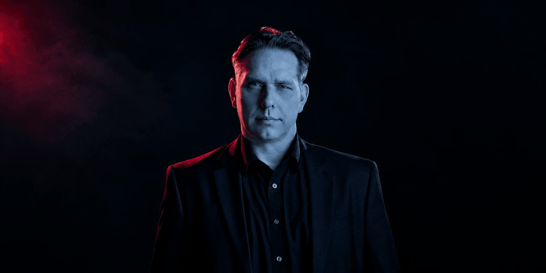 A man in a dark suit stands against a smoky, dramatic background.
