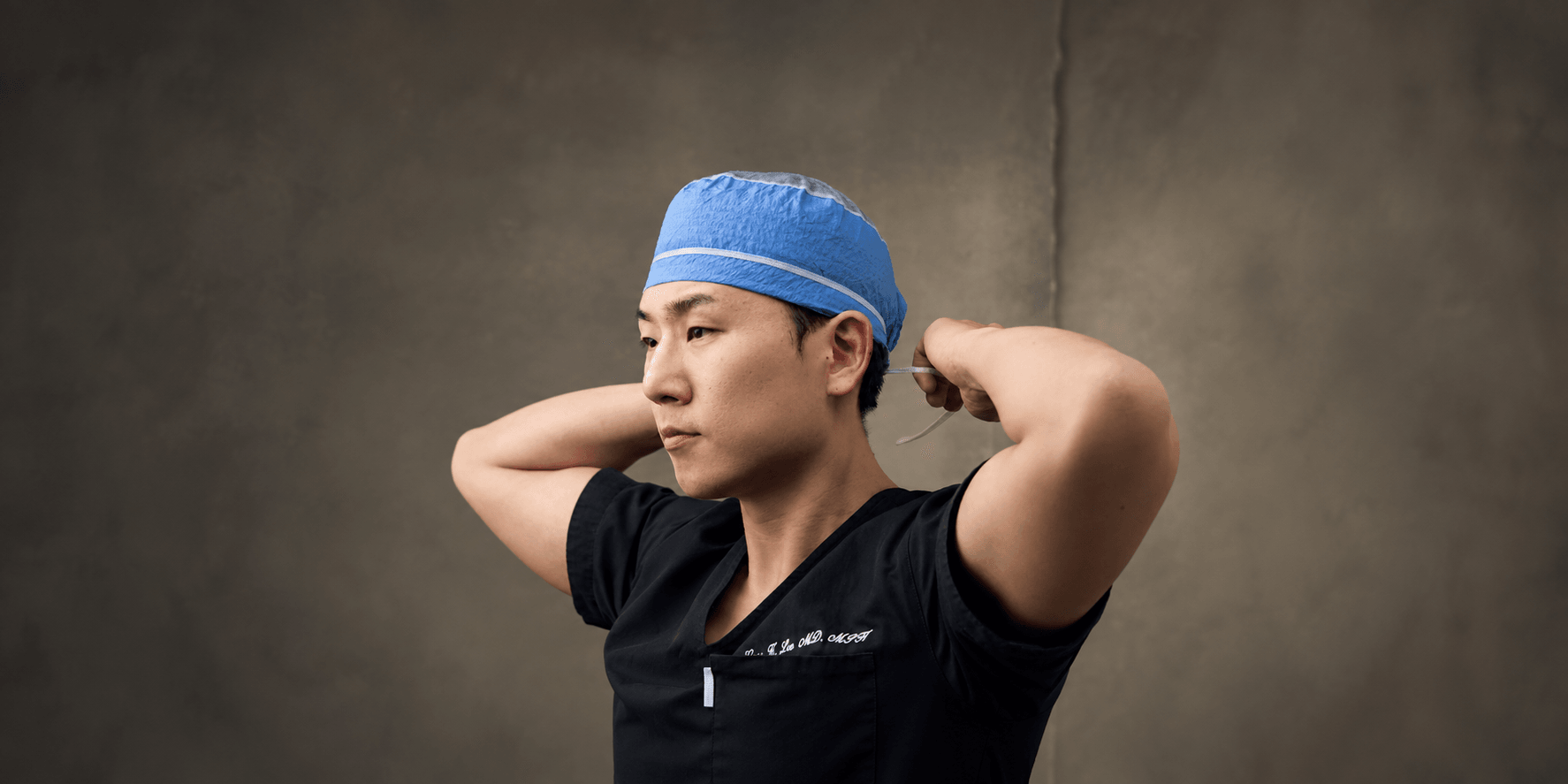 Surgeon adjusting his surgical cap, wearing black scrubs against a textured background