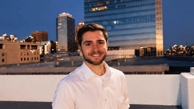 Smiling man in a white shirt against a city skyline at dusk