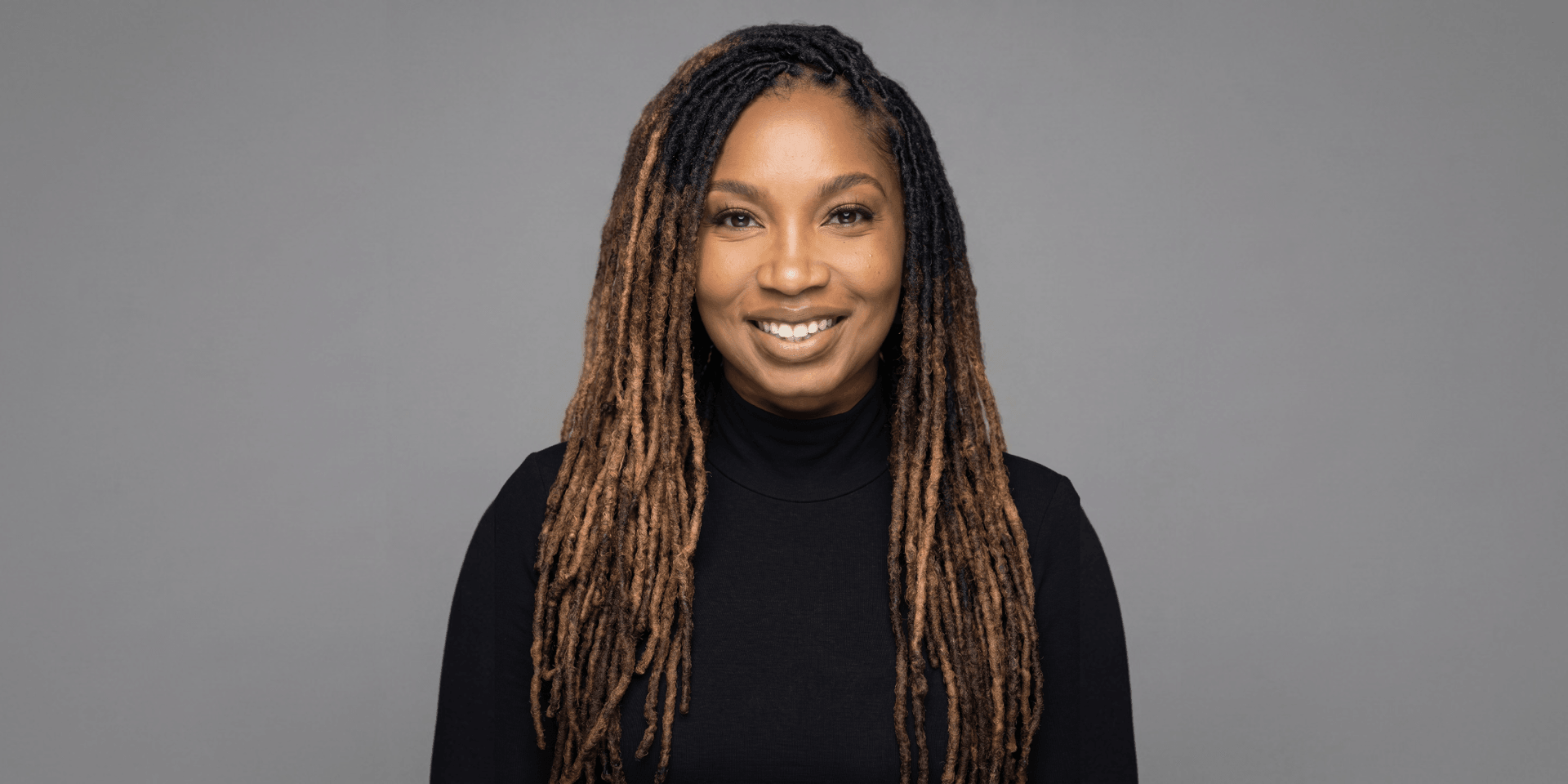 Smiling woman with long, styled hair wearing a black turtleneck against a gray background