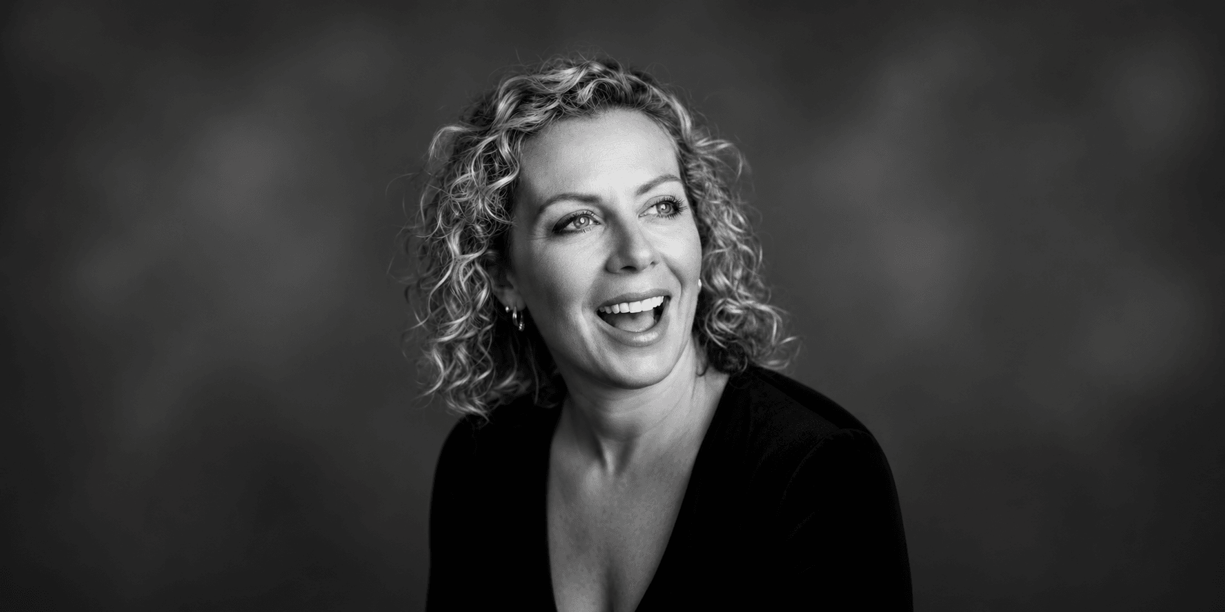 Woman with curly hair smiling, wearing a black top against a blurred background