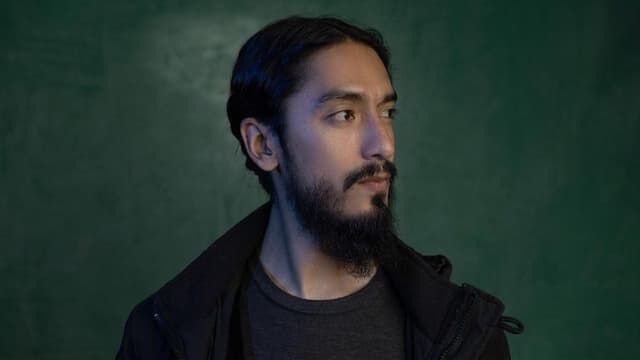 A man with long black hair and a beard poses against a green background