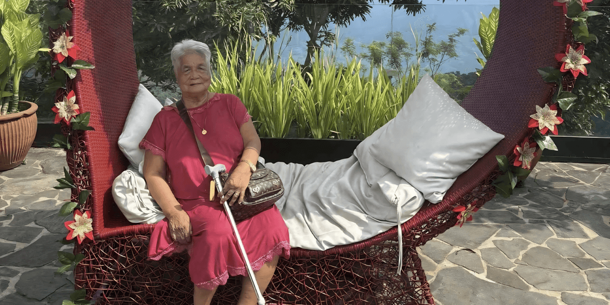 An elderly woman in a pink dress sits on a large woven hammock.