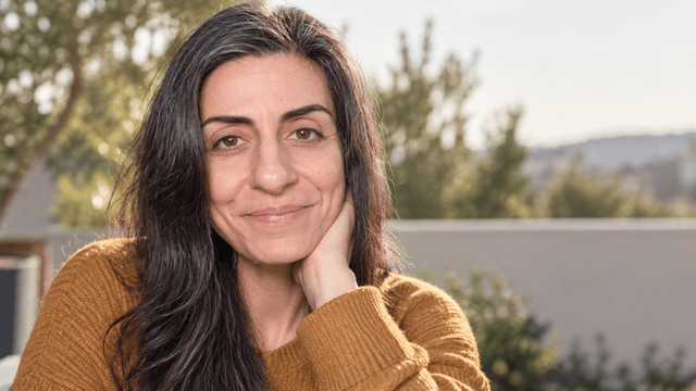 A woman with long dark hair smiles, resting her chin on her hand outdoors.