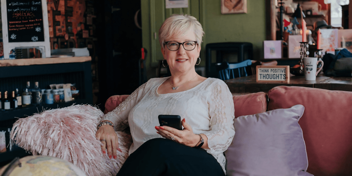 A woman with short blonde hair sits on a pink couch, smiling and holding a phone