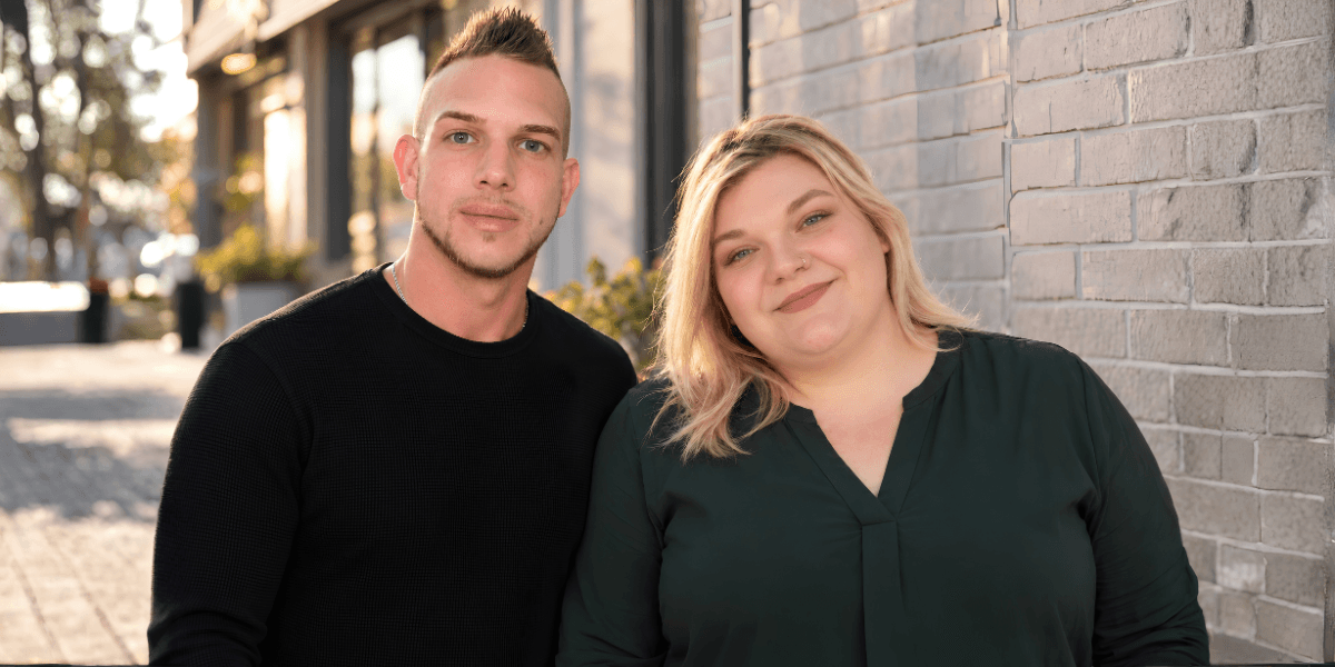 A young man and woman stand outdoors, smiling against a brick wall backdrop