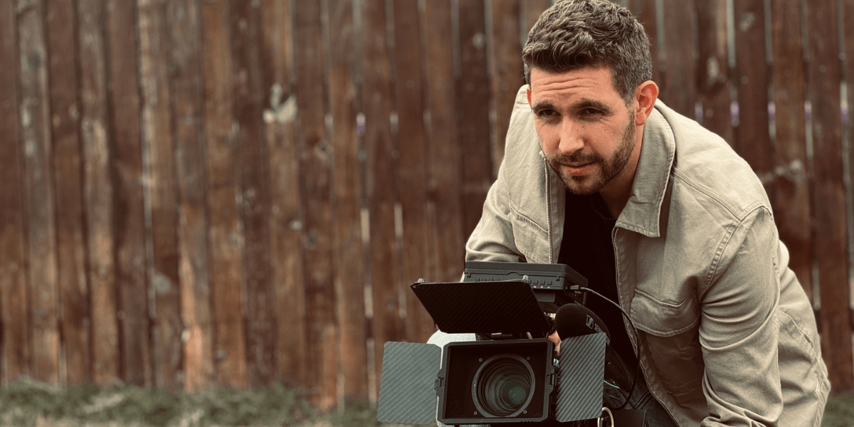 Man crouching with a camera in front of a wooden fence outdoors