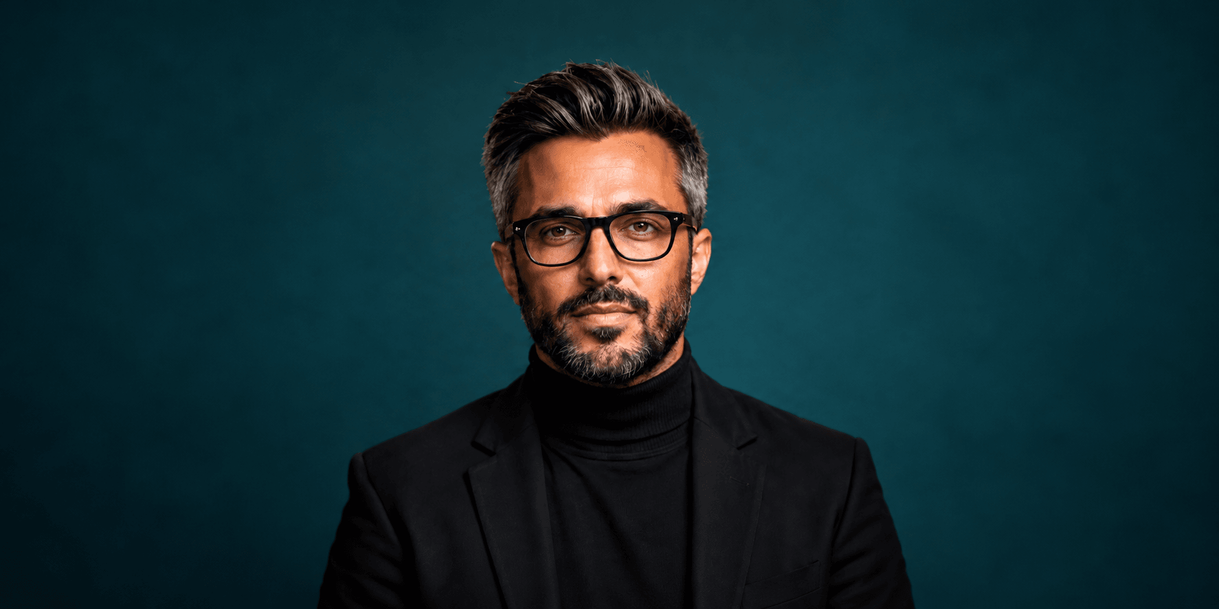 A man with glasses and a beard, wearing a black turtleneck and blazer, against a blue background