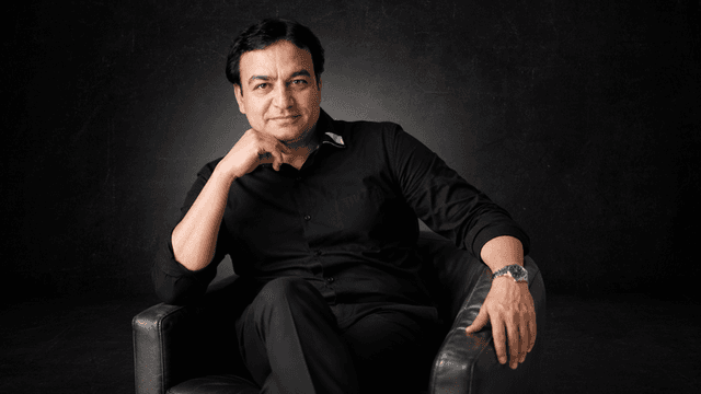 Man in a black shirt seated in a brown armchair against a dark background