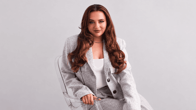 A woman in a gray suit sits confidently on a transparent chair against a light background