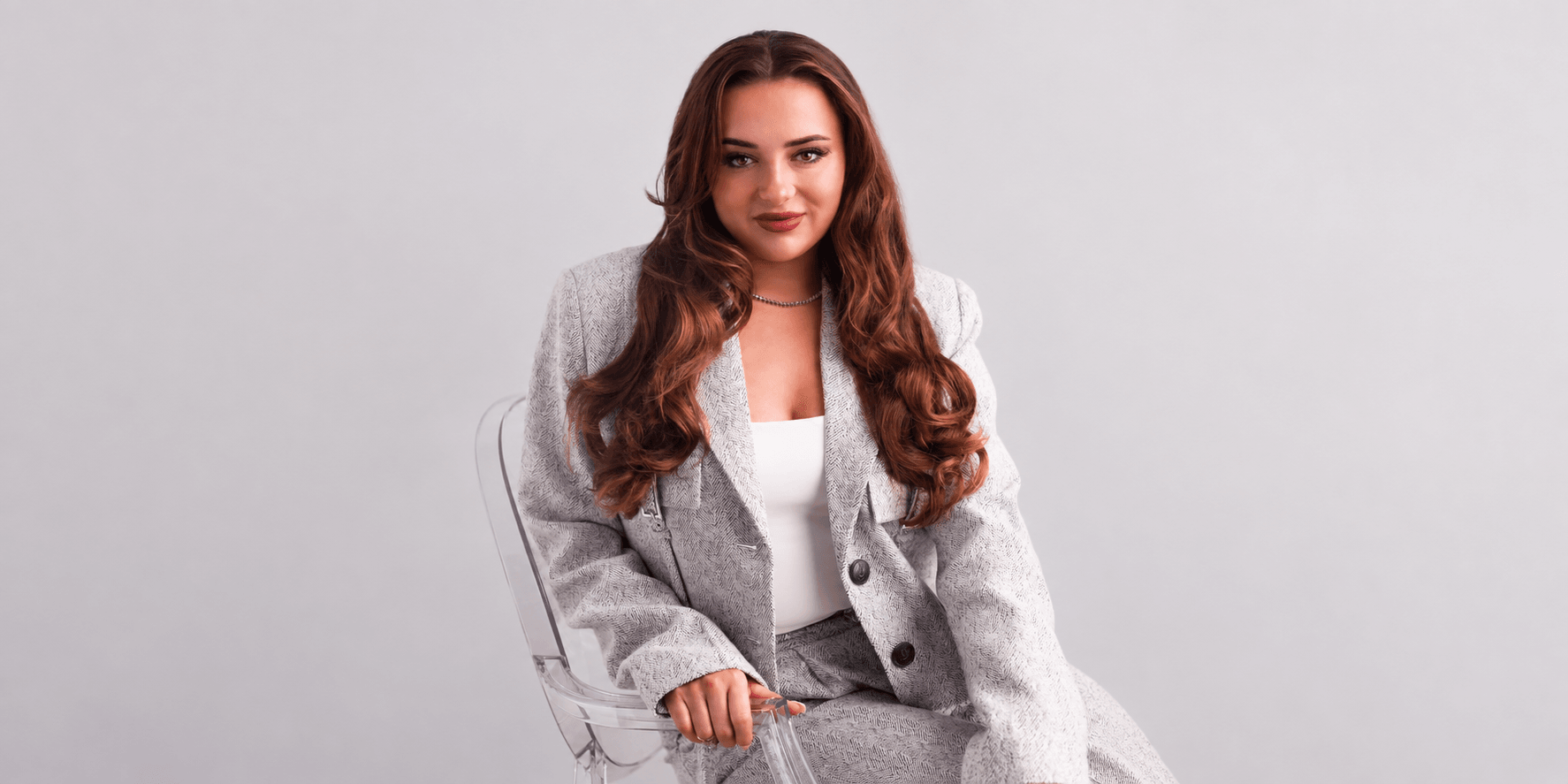 A woman in a gray suit sits confidently on a transparent chair against a light background