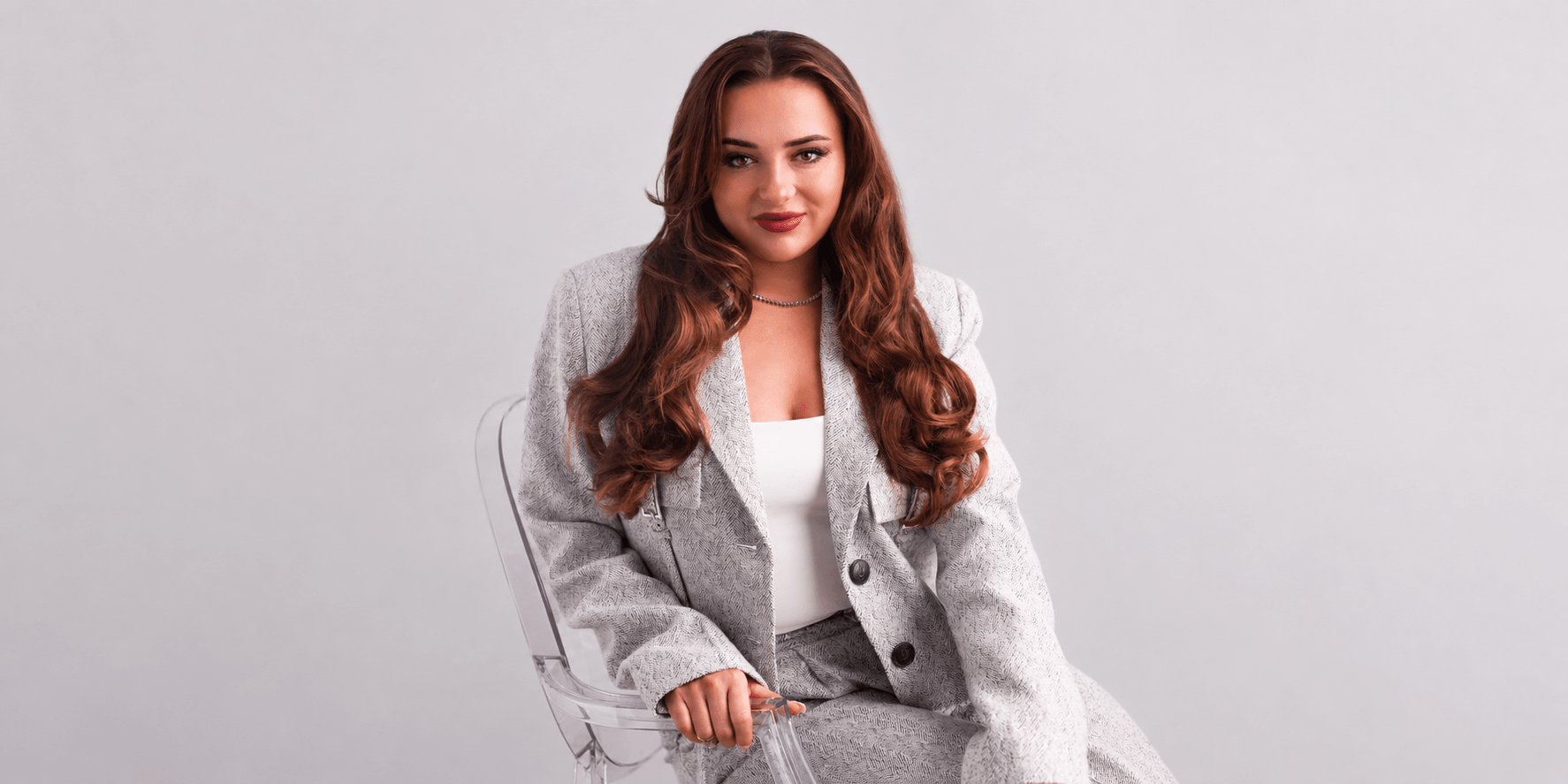 A woman in a gray suit sits confidently on a transparent chair against a light background