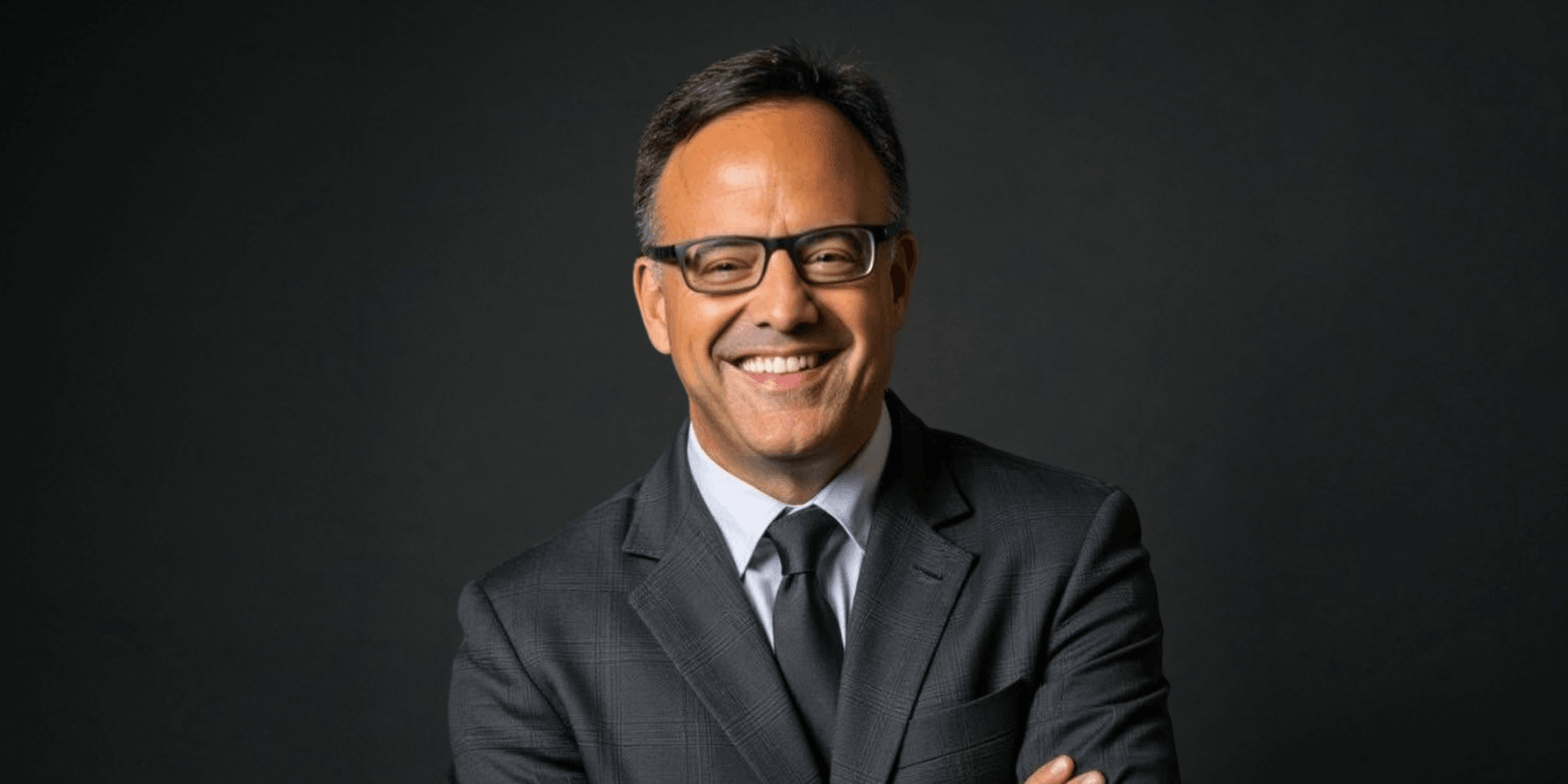 Smiling man in a suit and glasses with arms crossed against a dark background