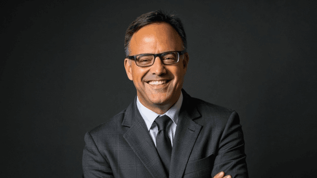 Smiling man in a suit and glasses with arms crossed against a dark background