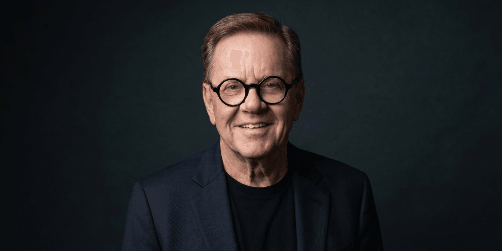 Smiling man with glasses wearing a blazer and black shirt against a dark background
