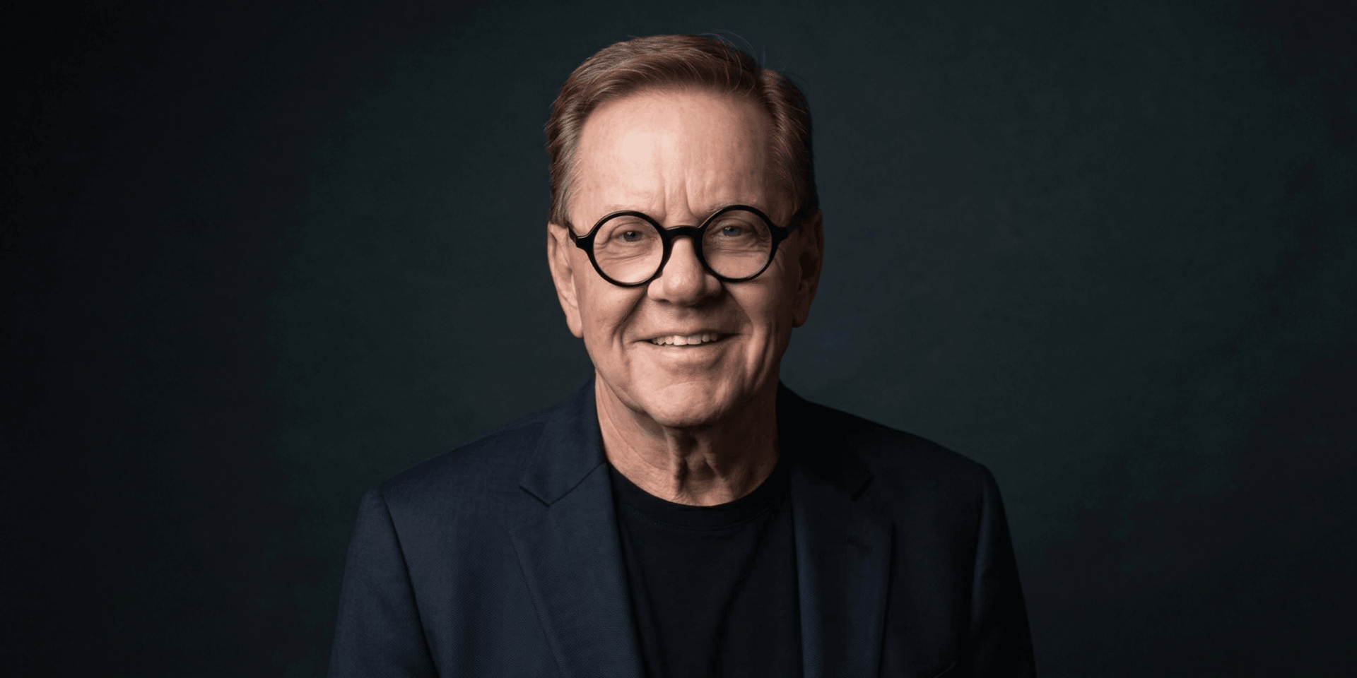 Smiling man with glasses wearing a blazer and black shirt against a dark background