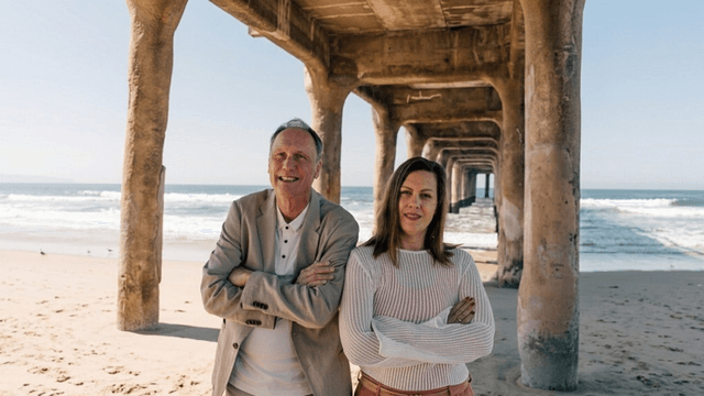 Two people smile under a pier by a beach, with waves in the background