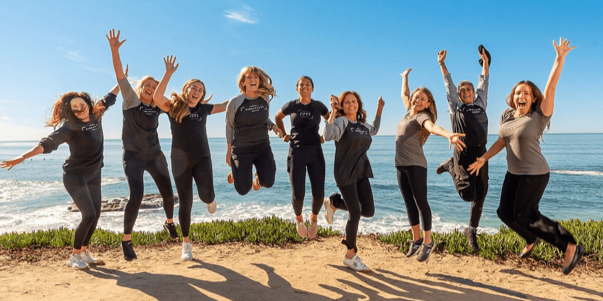 A group of eight people jumping joyfully by the ocean on a sunny day