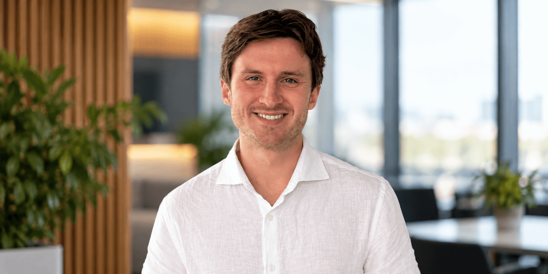 Smiling man in a white shirt standing in a modern office environment with plants.