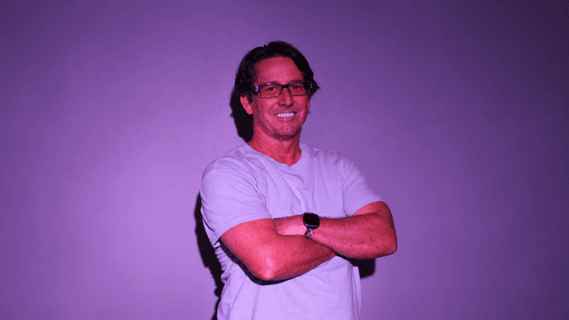 Smiling man in a light gray shirt stands with arms crossed against a purple backdrop