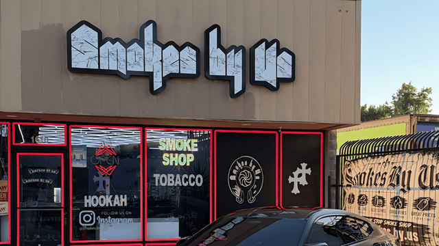 Storefront of a smoke shop with "Smokes By Us" sign and glass windows.