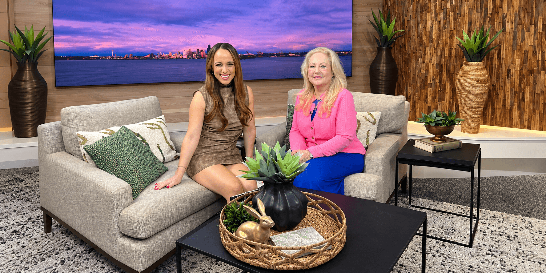 Two women seated on a couch in a modern studio with a city skyline backdrop