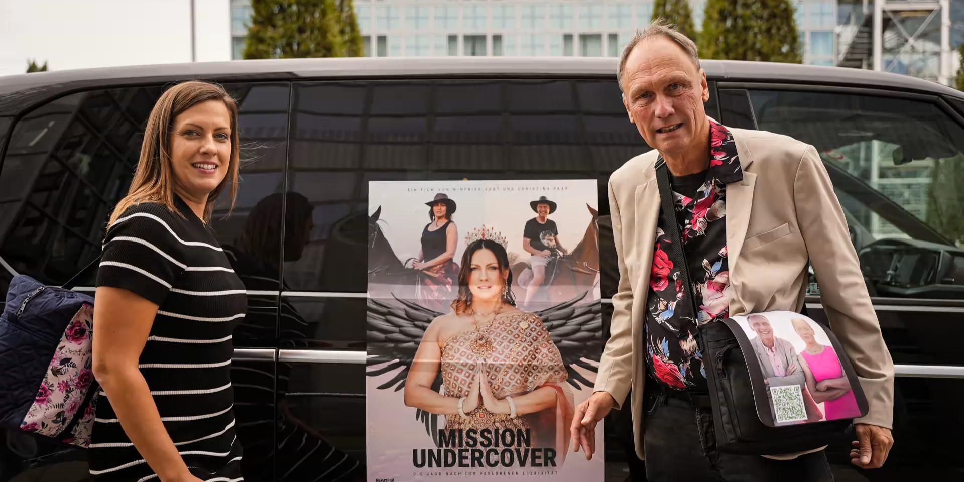 Two people posing beside a promotional poster for "Mission Undercover" near a vehicle