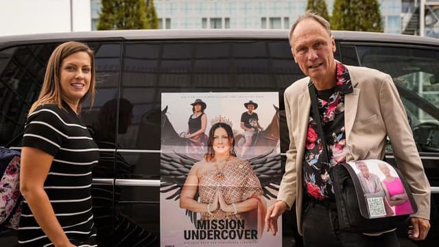Two people posing beside a promotional poster for "Mission Undercover" near a vehicle