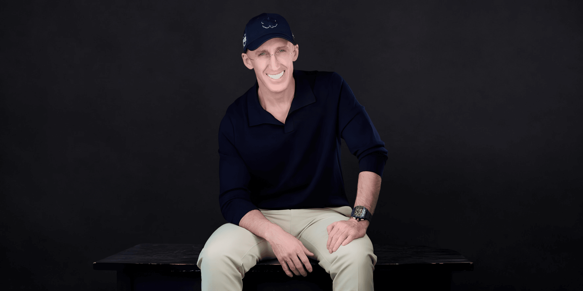 Man sitting on a bench, wearing a navy shirt and cap, smiling at the camera