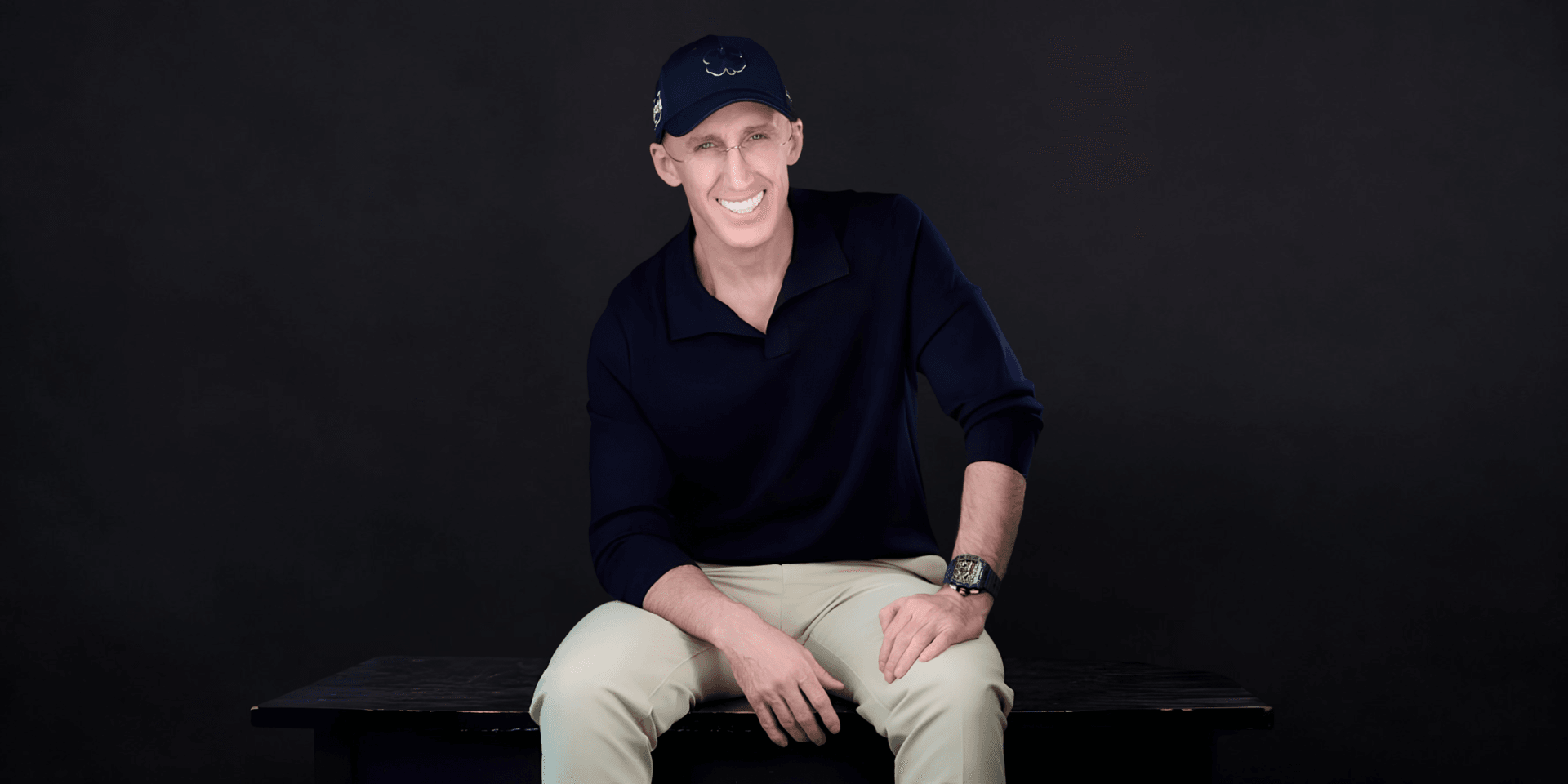 Man sitting on a bench, wearing a navy shirt and cap, smiling at the camera