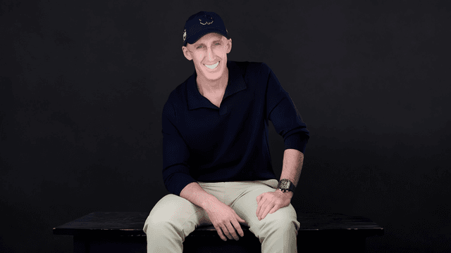 Man sitting on a bench, wearing a navy shirt and cap, smiling at the camera