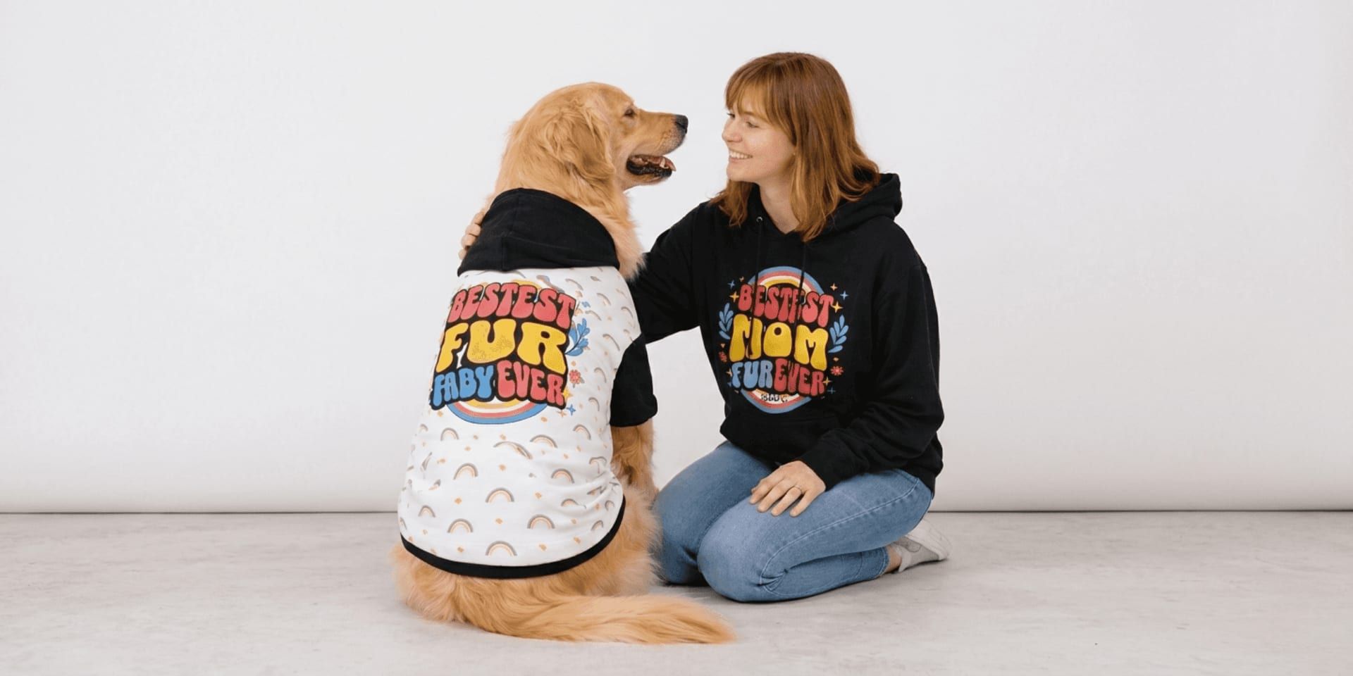 Woman and golden retriever wearing matching colorful hoodies, smiling together in a studio setting.