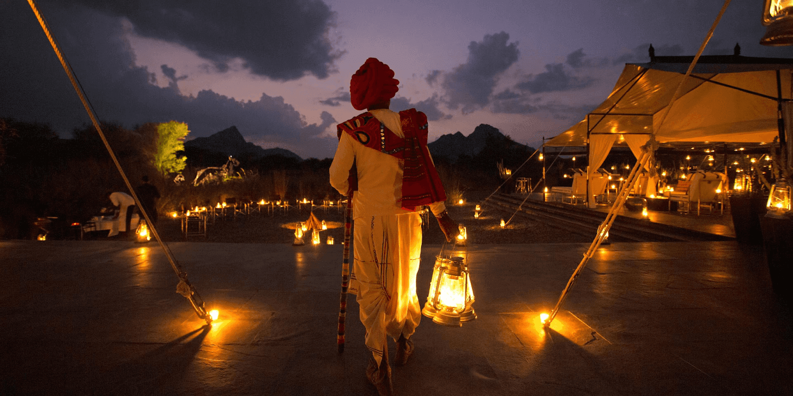 "A person in traditional attire holding a lantern walks through a candle-lit outdoor event at dusk, with mountains in the background and elegant tents set up for a luxurious experience."