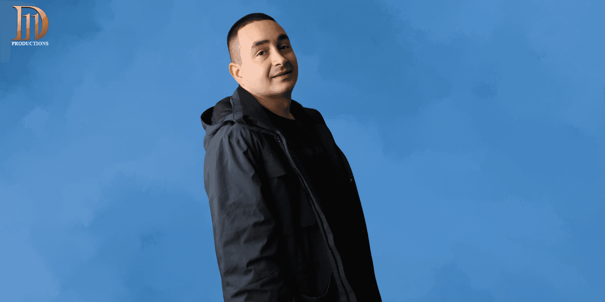 Portrait of a man in a dark jacket standing against a blue background, with the logo of JD Productions in the top left corner.
